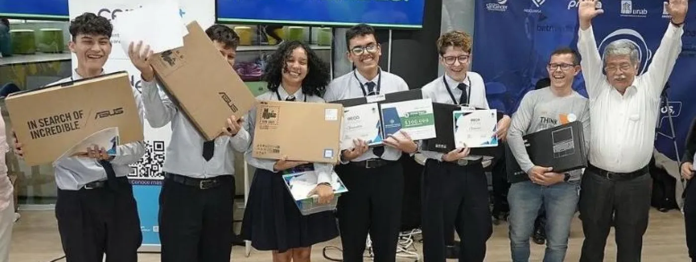 Grupo de estudiantes en uniforme escolar sosteniendo cajas de laptops con expresiones de alegría en un evento de entrega de equipos tecnológicos en un concurso en santander