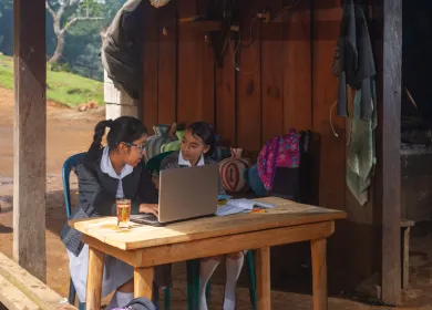 Dos niñas con uniforme escolar trabajando juntas frente a una laptop en un entorno rural, con mochilas y cántaros decorativos en el fondo, dentro de una estructura de madera.
